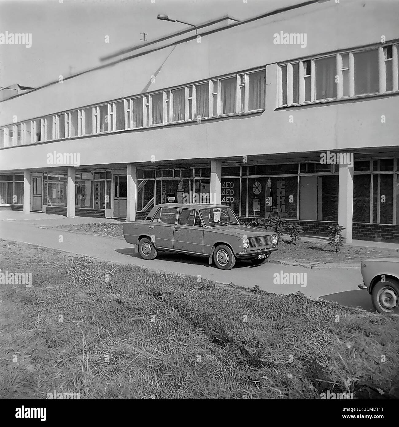 Une scène urbaine typique des dernières années de l'URSS est capturée dans cette photographie d'archives en noir et blanc de 1987 à Sloviansk, en RSS d'Ukraine. Une berline soviétique emblématique, la VAZ-2106 'Zhiguli', symbole de modeste prospérité à cette époque, est garée devant le restaurant 'Vesna' (printemps) dans le quartier résidentiel d'Artyom. Cette image sert de document historique précieux du mode de vie soviétique, des transports et de l'environnement urbain pendant la période charnière de la Perestroïka, mettant en valeur la vie quotidienne dans le contexte de l'architecture fonctionnaliste de l'époque Banque D'Images