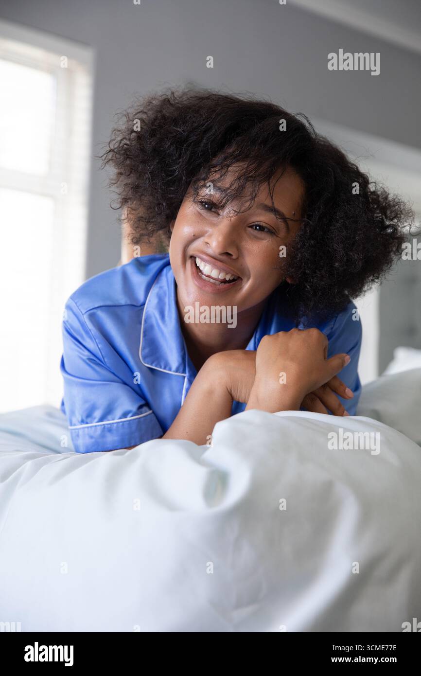 Femme afro-américaine s'inclinant sur le lit de la chambre en pyjama bleu profitant de la lumière à travers les stores Banque D'Images