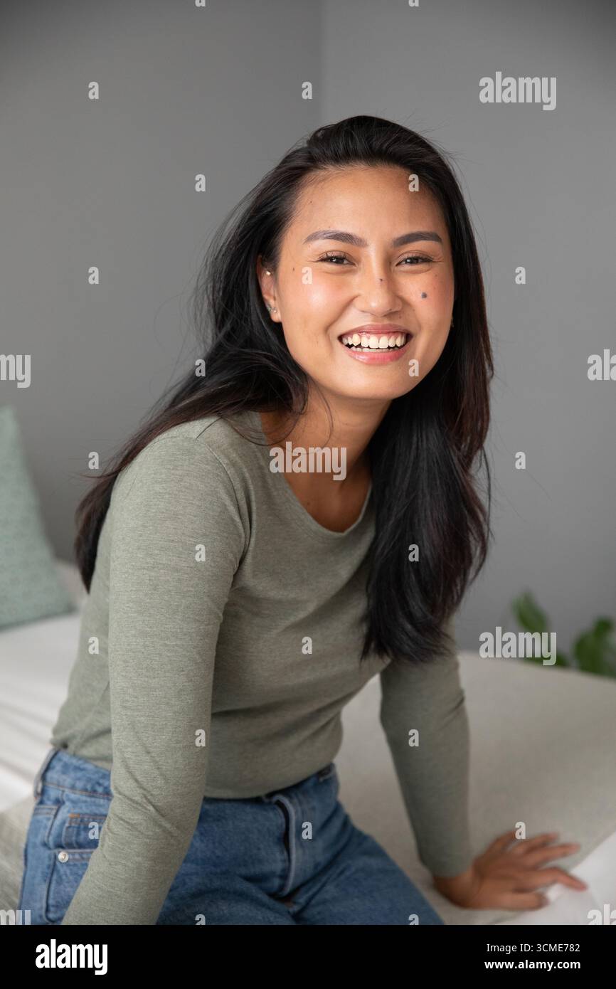 Femme asiatique assise sur un canapé dans la chambre portant un haut vert olive et un Jean avec coussin, plante Banque D'Images