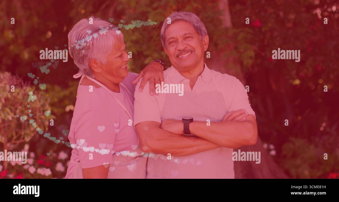 Posant couple senior portant tabliers avec smartwatch dans le jardin parmi les arbustes à fleurs, lumières de coeur Banque D'Images