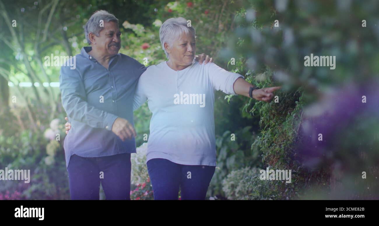 Flâner un couple de retraités bras dans bras, pointant vers le feuillage le long d'un chemin de jardin ensoleillé Banque D'Images