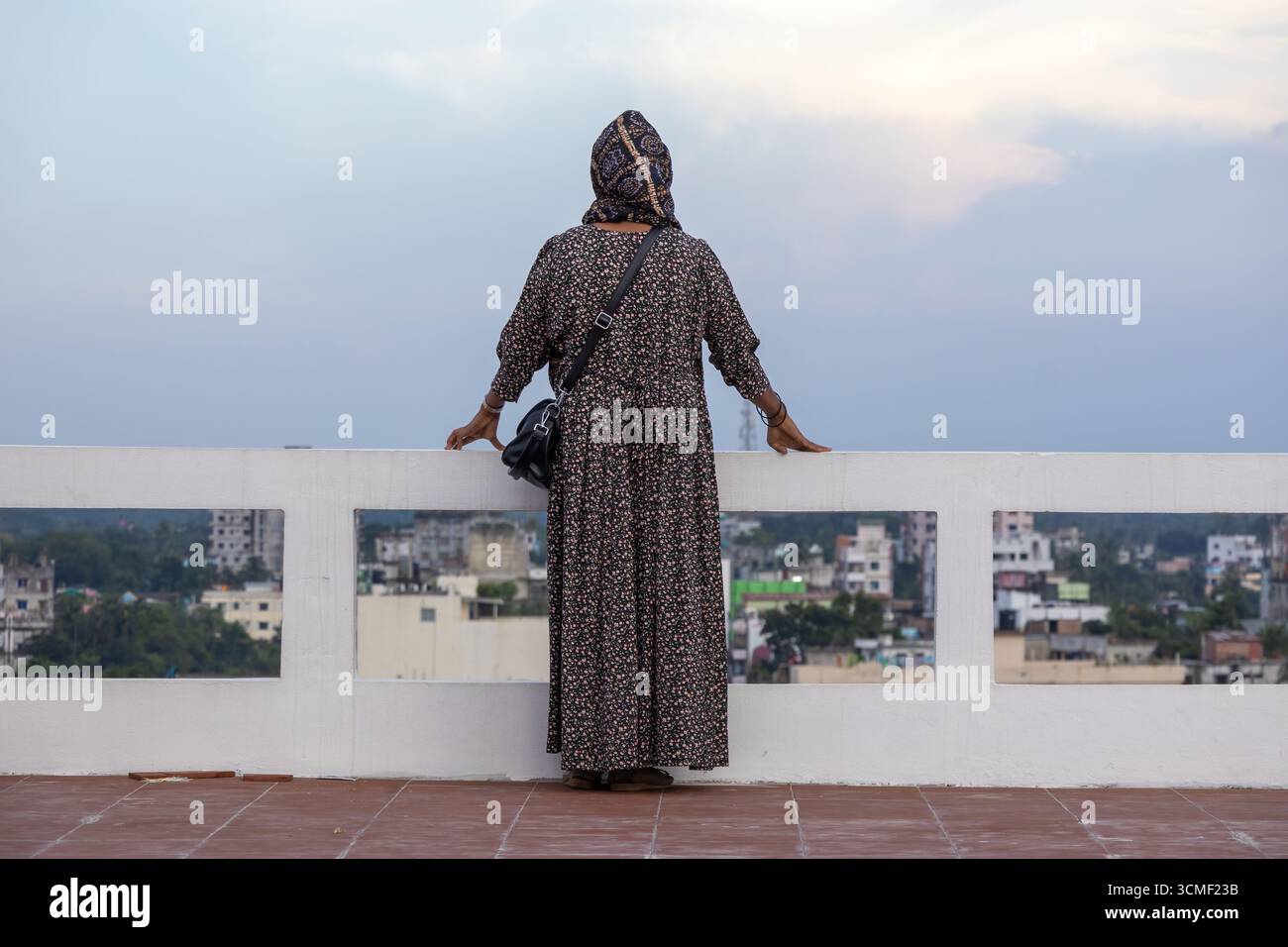 Vue arrière d'une femme musulmane bangladaise dans une abaya florale et hijab, debout sur le toit d'un bâtiment, regardant la ville et le paysage naturel. Banque D'Images