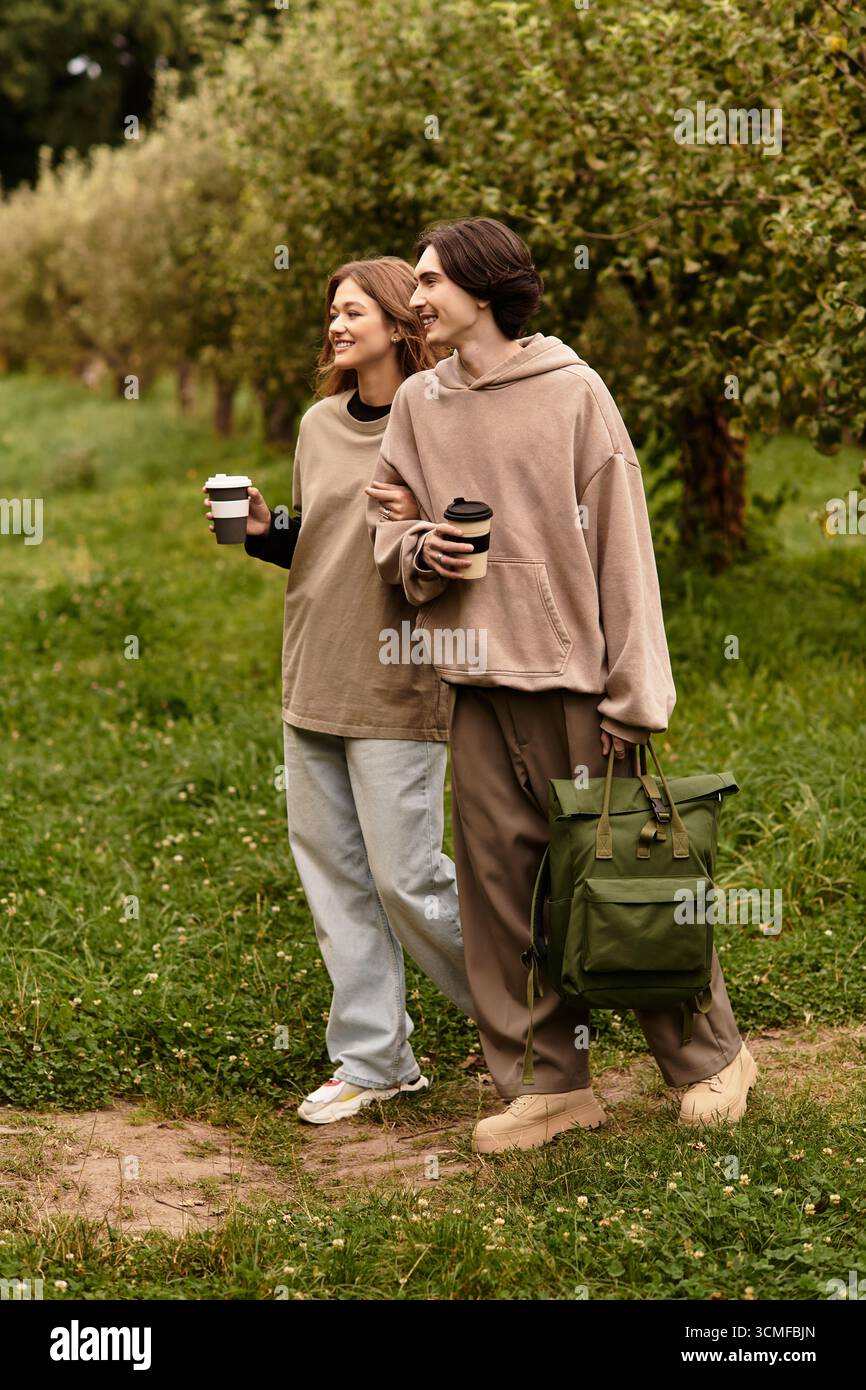 Deux amoureux se promènent main dans la main dans un verger luxuriant, partageant boissons chaudes et sourires. Banque D'Images