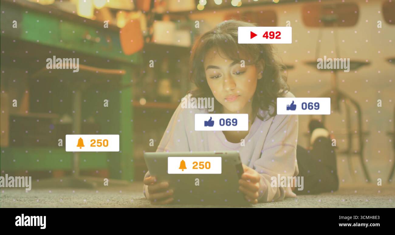 Femme adulte couchée tenant la tablette sur la moquette de la maison, avec des superpositions d'icône de notification et des lumières de chaîne Banque D'Images