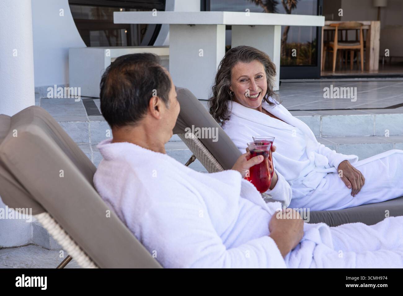 Couple de personnes âgées diversifié assis sur des chaises longues dans le patio du spa en sirotant une boisson dans le pichet en verre Banque D'Images