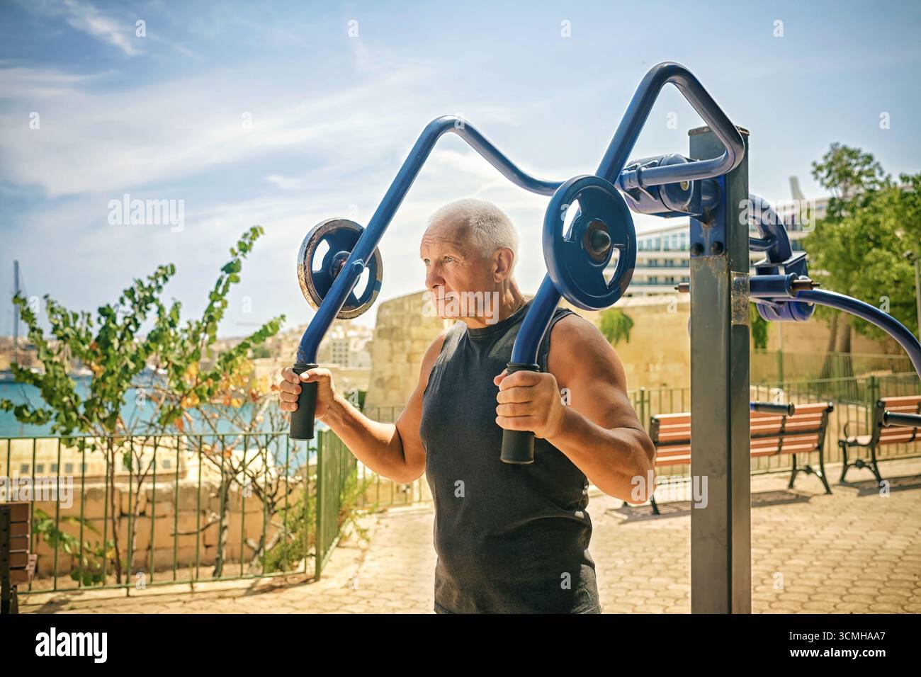 Homme âgé actif avec les cheveux gris dans le débardeur noir exercice sur l'équipement de fitness extérieur par la mer sous le ciel ensoleillé. Banque D'Images
