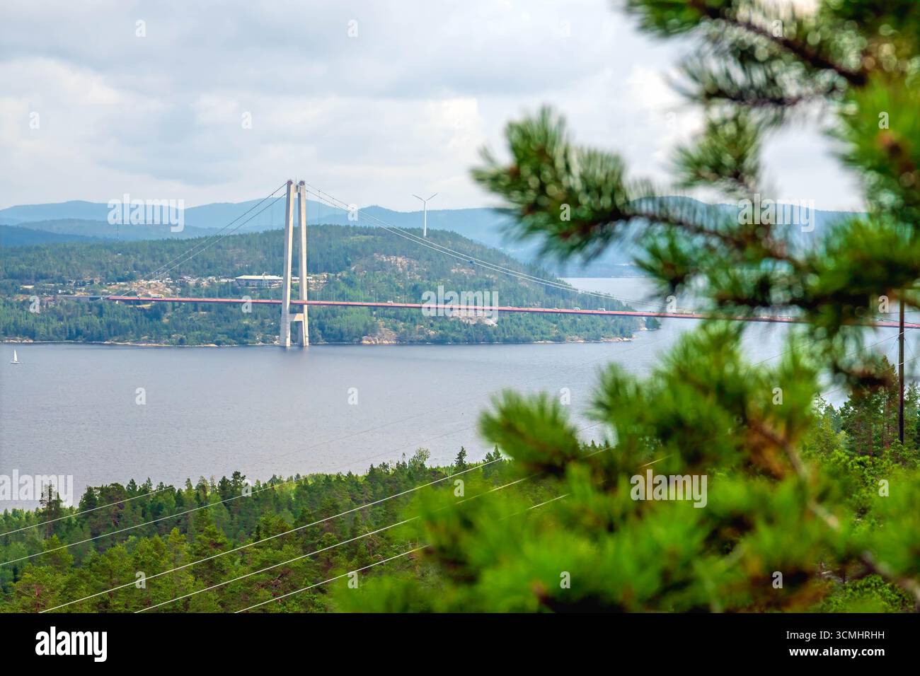High Coast Bridge, une structure à haubans de câbles en Suède, enjambe une étendue d'eau, reliant des rivages sous un ciel nuageux, avec des éoliennes lointaines et f Banque D'Images