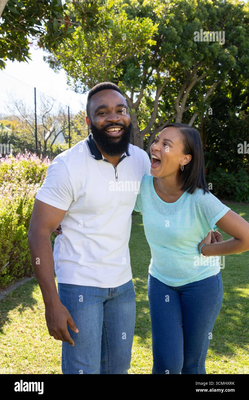 Couple diversifié debout dans le jardin ensoleillé sur la pelouse herbeuse penché l'un vers l'autre riant Banque D'Images