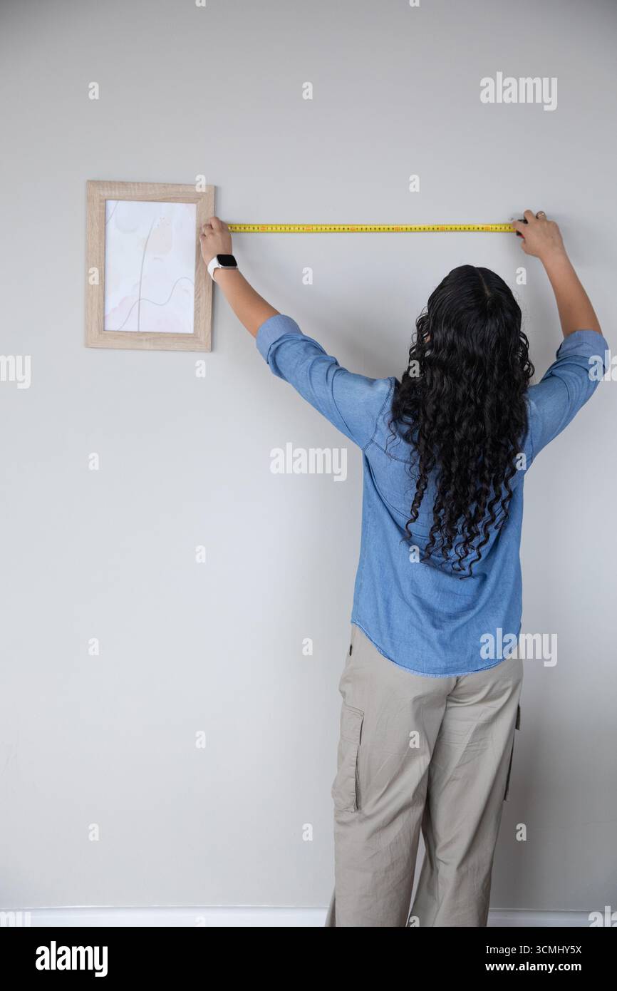 Femme afro-américaine mesurant le mur gris à la maison à l'aide d'un mètre ruban à côté de l'impression d'art encadrée Banque D'Images Femme afro-américaine mesurant le mur gris à la maison à l'aide d'un mètre ruban à côté de l'impression d'art encadrée Banque D'Images