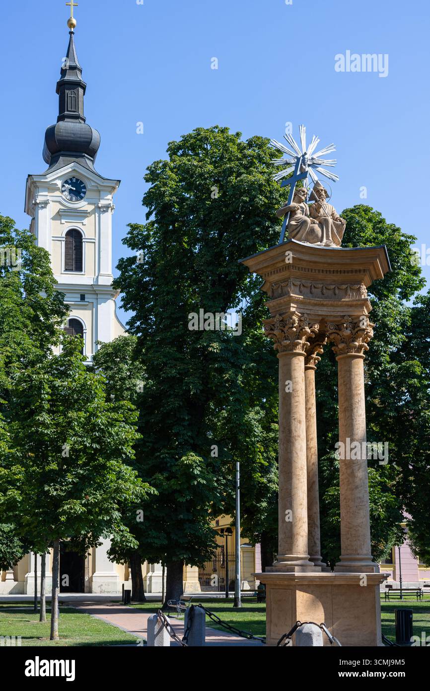 Une vue panoramique d'une tour d'église historique avec une horloge, accompagnée d'une statue de piédestal détaillée, entourée d'arbres verts luxuriants sous un bleu vif s. Banque D'Images