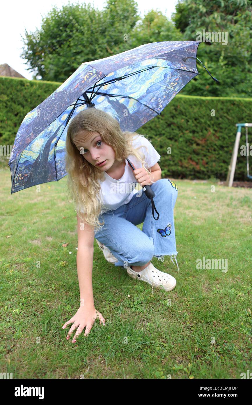 Fille avec un parapluie coloré dans le jardin regardant un contact direct des yeux accroupissant sur le sol touchant l'herbe avec la main Banque D'Images