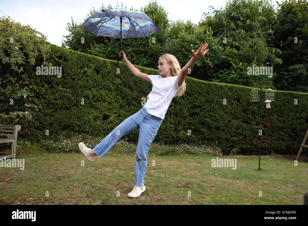Fille avec un parapluie coloré dans le jardin donnant un coup de pied haut de jambe portant un Jean et un t-shirt Banque D'Images
