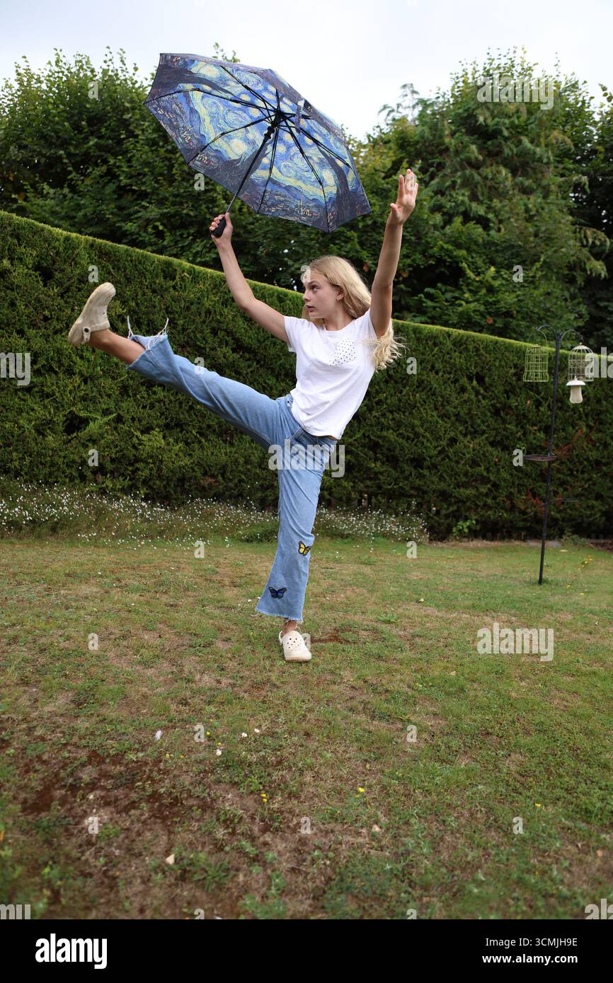 Fille avec un parapluie coloré dans le jardin donnant un coup de pied haut de jambe portant un Jean et un t-shirt équilibre équilibre gymnaste Banque D'Images