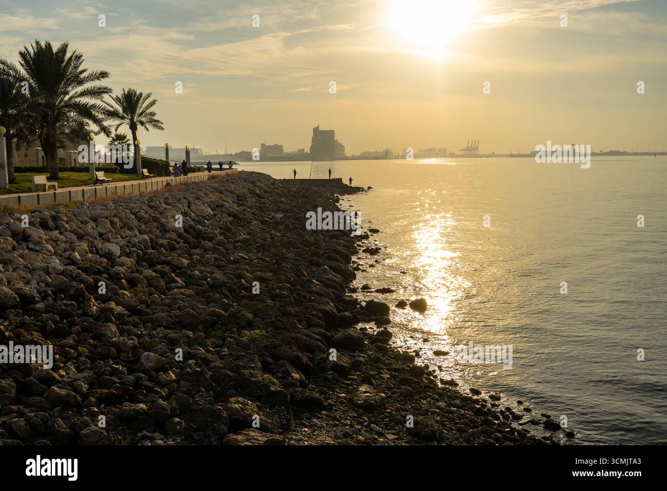 Lever de soleil doré sur un rivage rocheux dans la ville de Koweït, avec les pêcheurs et les piétons profitant de la matinée paisible au bord de l'eau. Banque D'Images