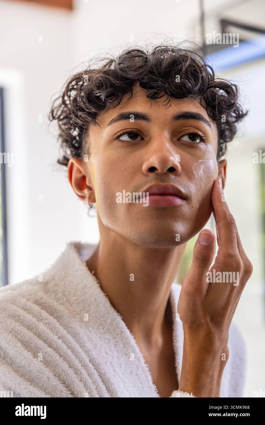 Homme appliquant la crème pour le visage, debout devant le miroir de la salle de bain portant peignoir et boucle d'oreille Banque D'Images