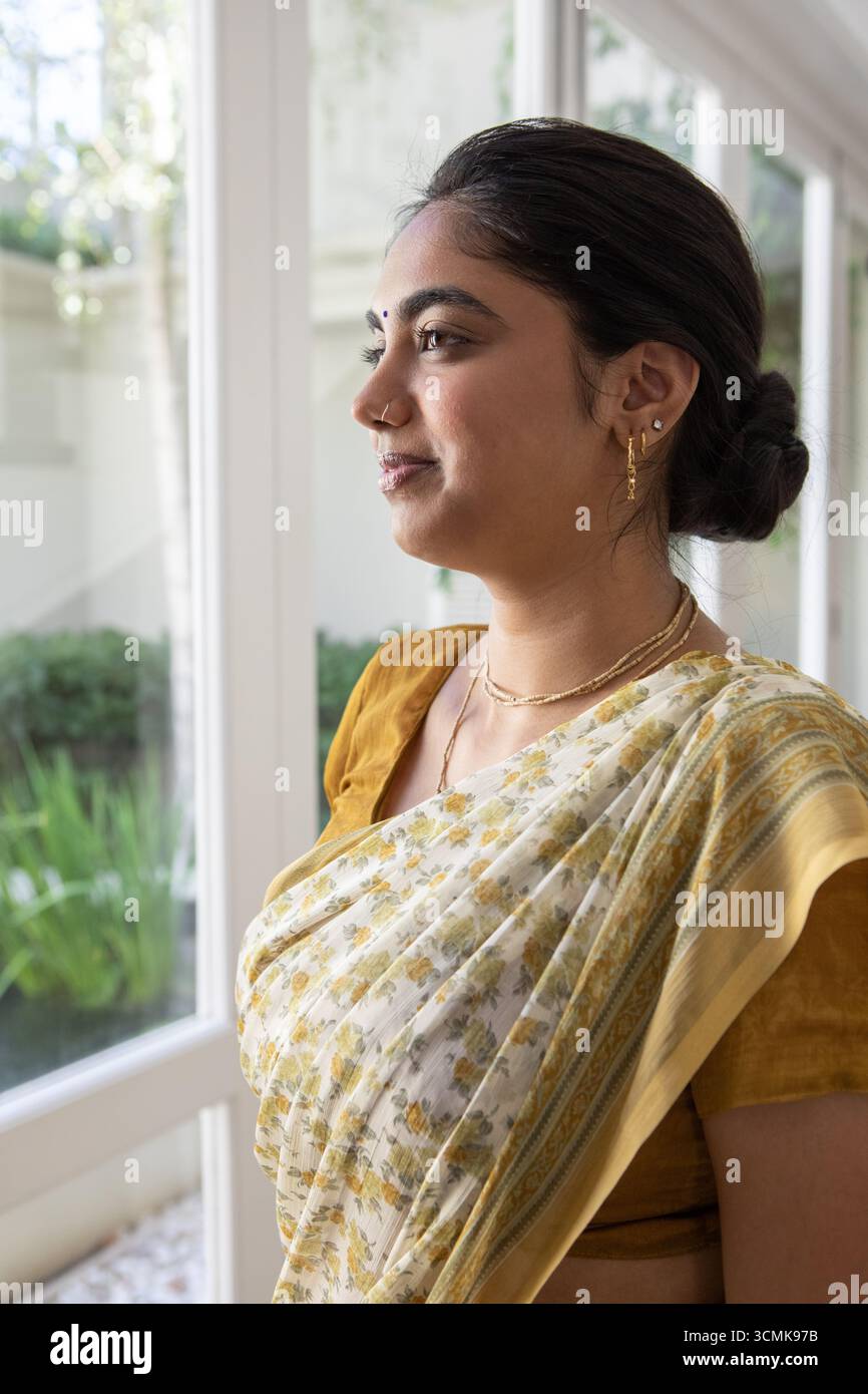 Femme indienne debout par des fenêtres en verre à la maison et regardant le jardin dans le saree floral Banque D'Images