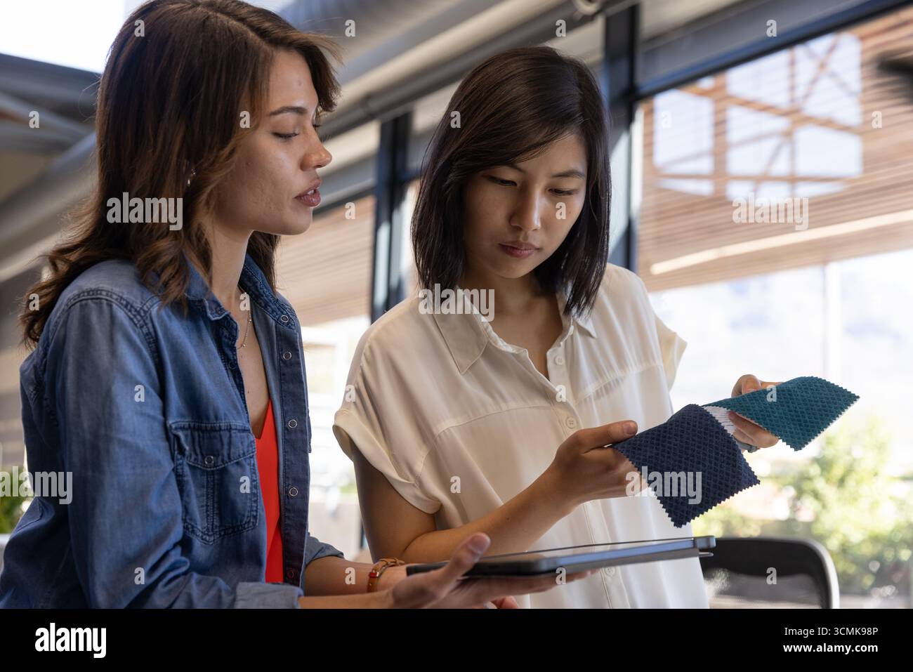 Diverses collègues féminines debout près des fenêtres, examinant des échantillons de tissu bleu avec tablette dans le studio Banque D'Images