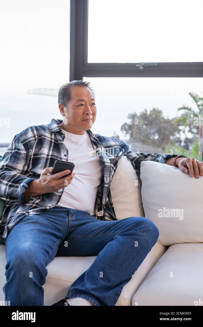 Homme chinois asiatique senior assis sur le canapé dans le salon tenant smartphone Banque D'Images