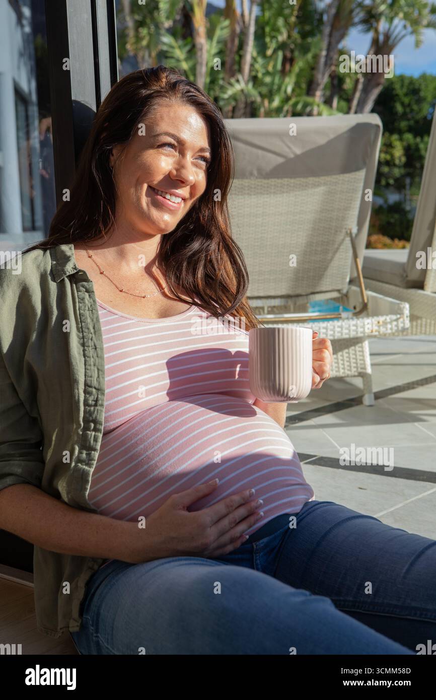 Femme enceinte appuyée contre la porte vitrée tenant une tasse à café sur un patio ensoleillé avec des meubles en osier Banque D'Images