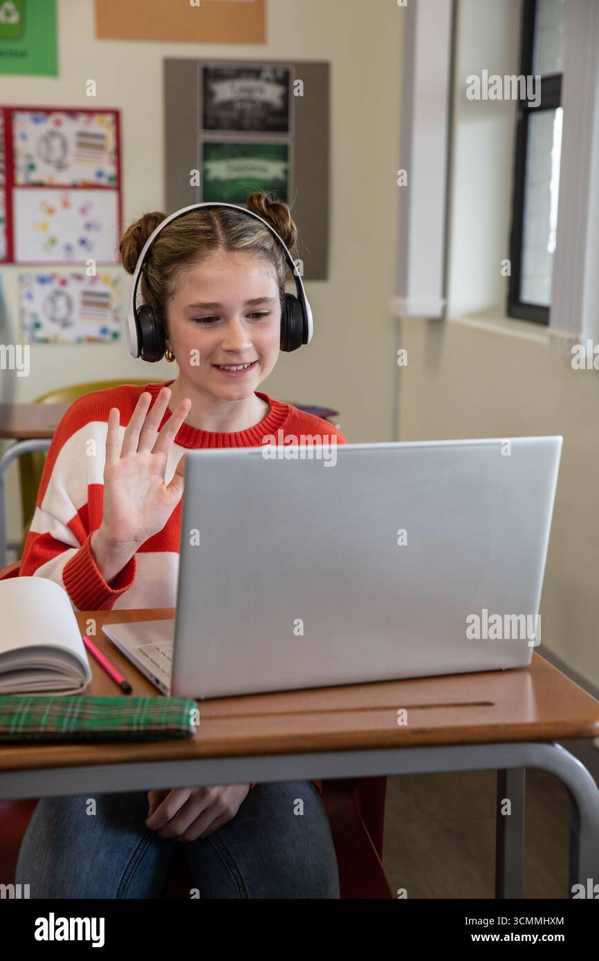 Jeune fille pré-adolescente assise au bureau dans la salle de classe dans un pull rayé à l'aide d'un ordinateur portable et d'un casque Banque D'Images