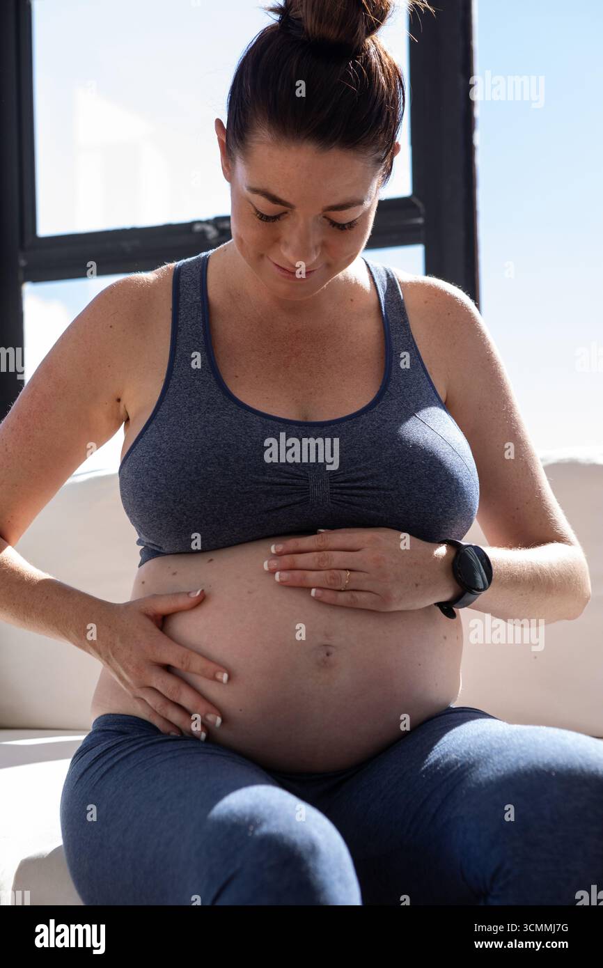 Femme adulte moyenne assise sur un canapé léger près de la fenêtre, berçant le ventre enceinte et montrant smartwatch Banque D'Images