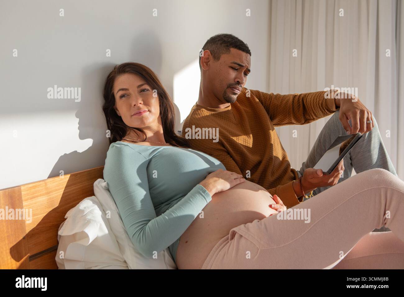 Couple diversifié reposant sur le lit dans la chambre avec des oreillers, tenant la tablette et caressant l'abdomen Banque D'Images