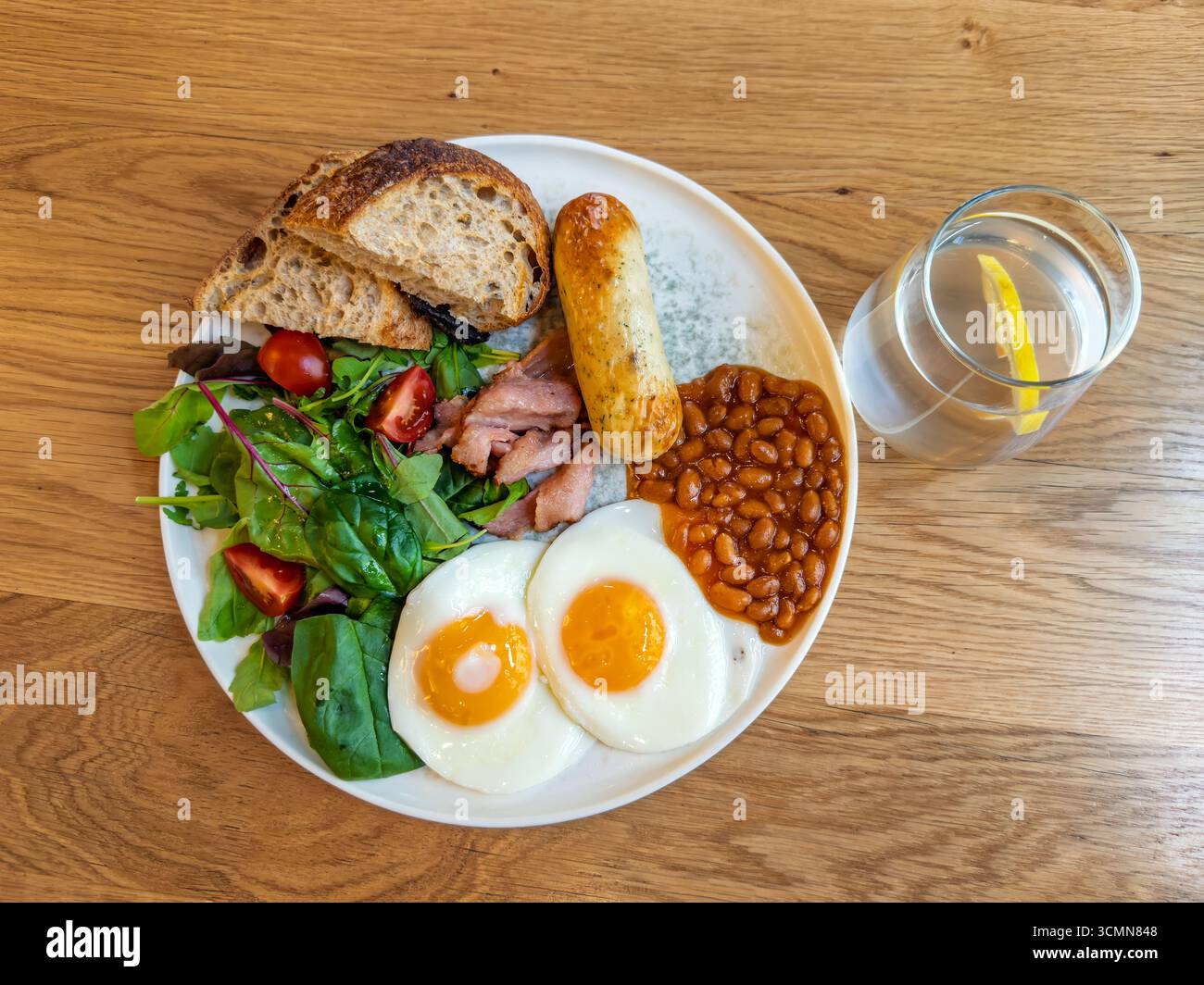 Petit déjeuner anglais complet avec œufs au plat, bacon, haricots, saucisses et salade fraîche servis sur une assiette blanche avec de l'eau, vue de dessus. Assiette de petit déjeuner copieux Banque D'Images