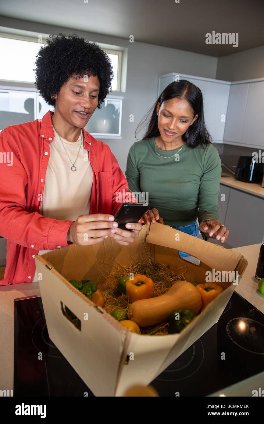 Couple afro-américain et indien déballant la boîte de produits à l'îlot de cuisine, examinant smartphone Banque D'Images Couple afro-américain et indien déballant la boîte de produits à l'îlot de cuisine, examinant smartphone Banque D'Images