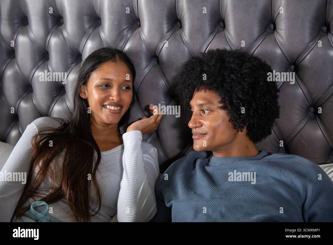 Couple diversifié parlant tout en étant allongé sur le lit dans la chambre, tête de lit grise touffetée, oreillers Banque D'Images