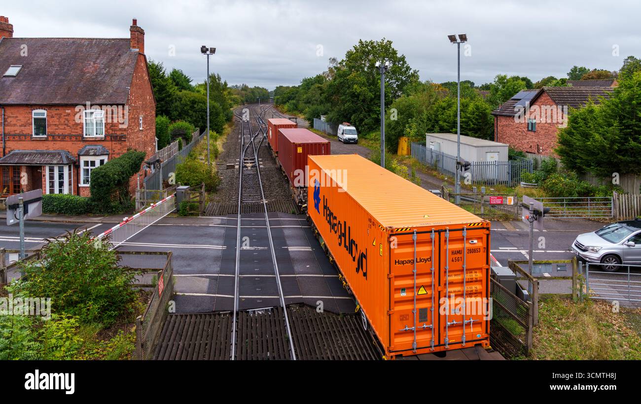 Un train de marchandises transportant des conteneurs intermodaux traverse un passage à niveau avec des barrières fermées, bloquant la circulation automobile Banque D'Images