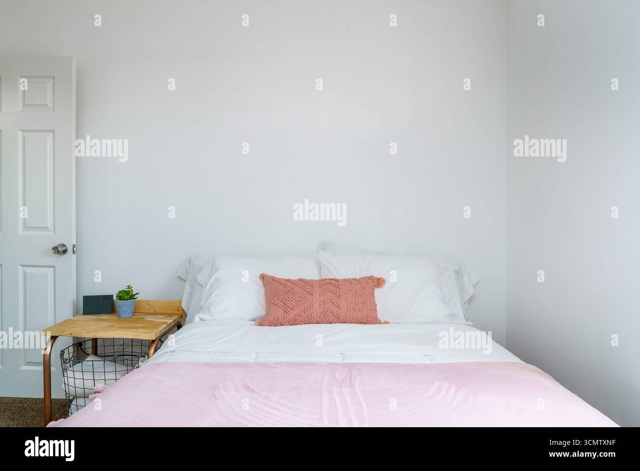 Chambre blanche avec oreiller chenille rose, couverture de plaid et table d'appoint avec panier. Vue de face. Banque D'Images