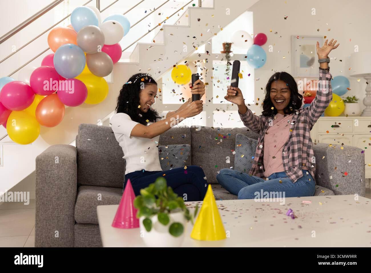Maman diversifiée et fille adolescente éclatant des canons à confettis sur un canapé gris dans le salon Banque D'Images