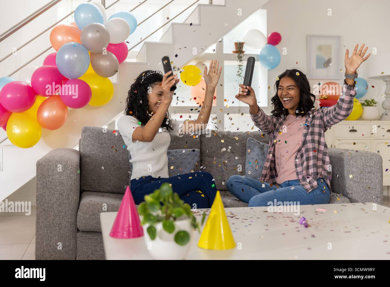 Maman diversifiée et fille adolescente assis sur le canapé à la maison, éclatant des poppers de confettis noirs Banque D'Images