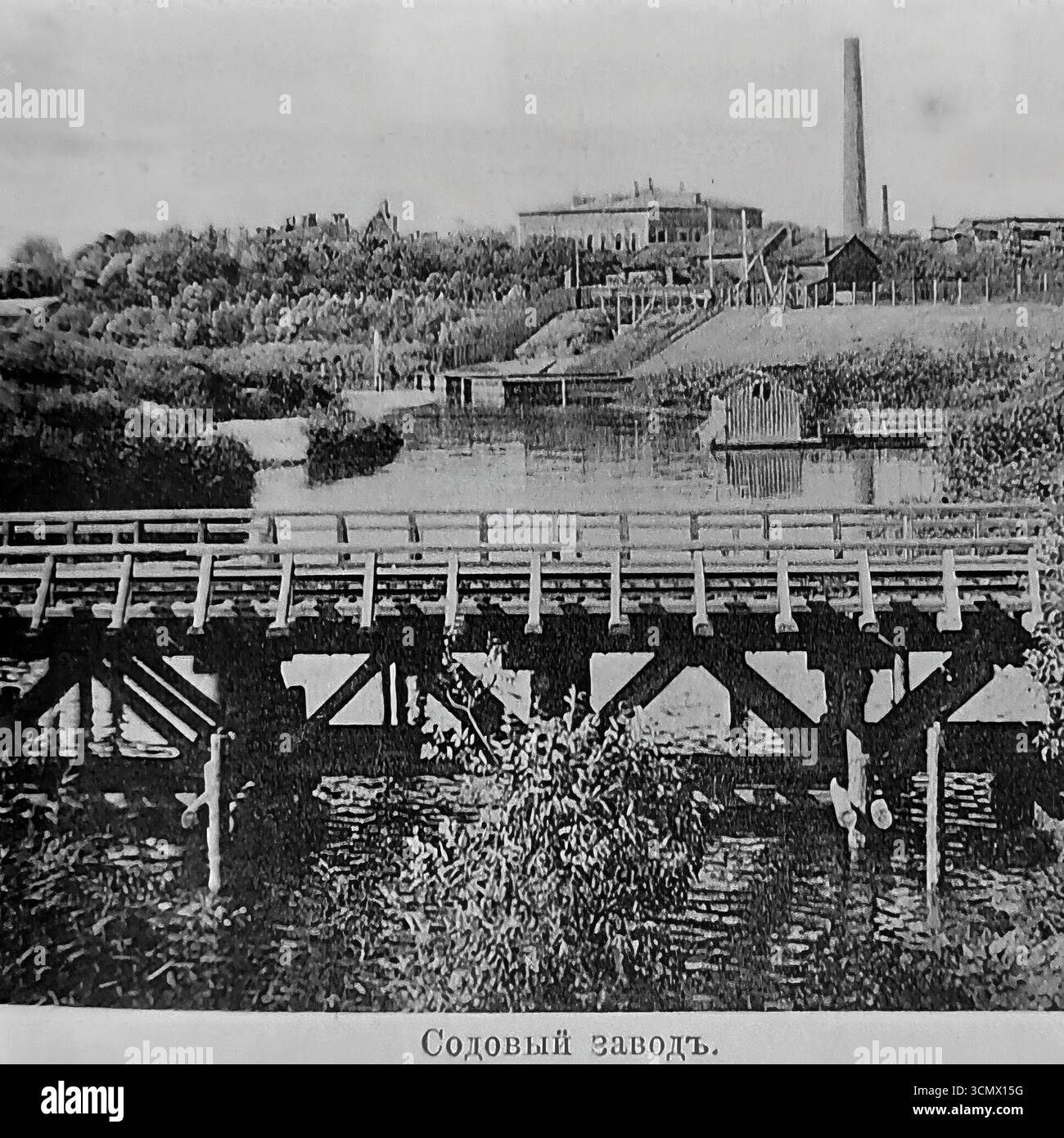 Une photographie historique des années 1900-1910, montrant l'usine de soude dans la ville de Sloviansk, dans l'Empire russe. L'énorme entreprise chimique, située sur les rives de la rivière Kazennyi Torets, était un pilier de l'industrie du Donbass et un symbole du développement industriel de la région. Au premier plan se trouve un pont en bois traversant la rivière contre le paysage industriel Banque D'Images