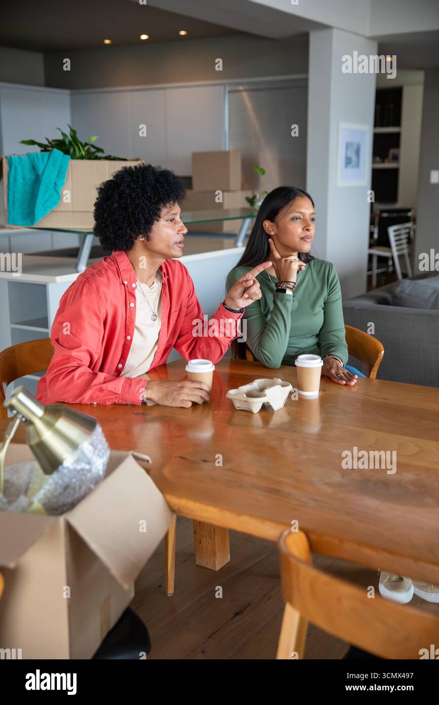 Couple diversifié assis et discutant se déplacer à la table à manger de l'appartement, tasses à café, boîtes de déménagement Banque D'Images Couple diversifié assis et discutant se déplacer à la table à manger de l'appartement, tasses à café, boîtes de déménagement Banque D'Images
