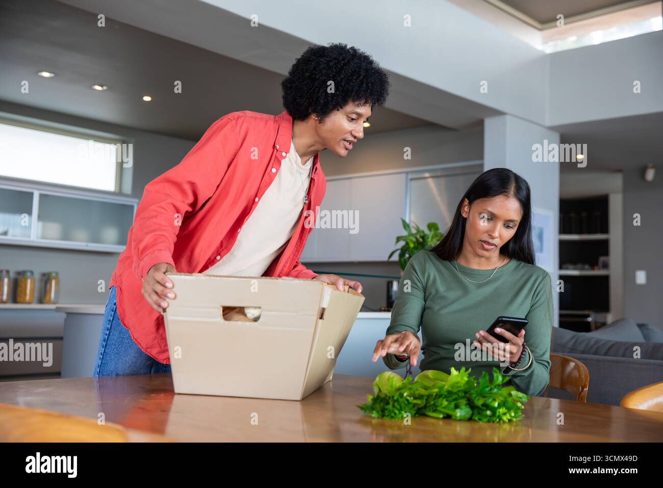 Couple diversifié déballer la boîte en carton et examiner les verts feuillus, en utilisant un smartphone dans la cuisine Banque D'Images