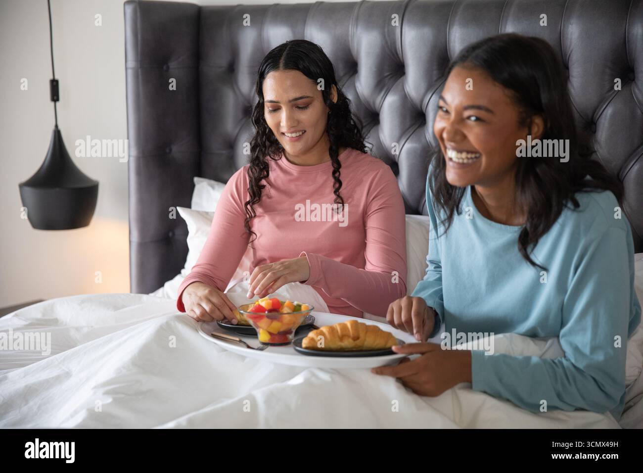 Diverses amies féminines assis sur le lit sous la lampe partageant des plateaux de petit déjeuner avec des fruits, des pâtisseries Banque D'Images Diverses amies féminines assis sur le lit sous la lampe partageant des plateaux de petit déjeuner avec des fruits, des pâtisseries Banque D'Images