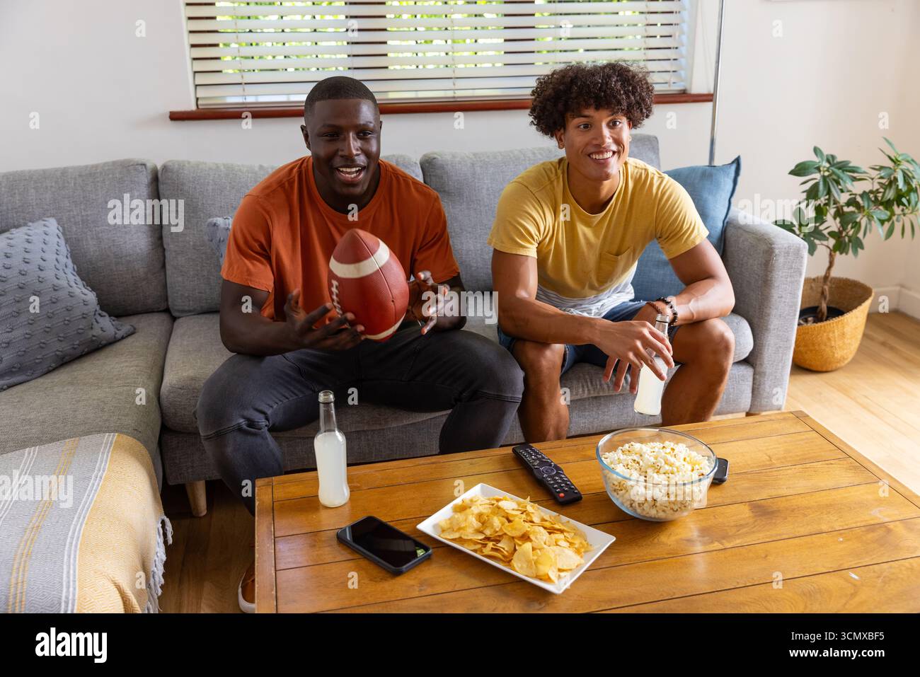 Divers amis masculins tenant le football américain, assis sur le canapé dans le salon avec pop-corn Banque D'Images