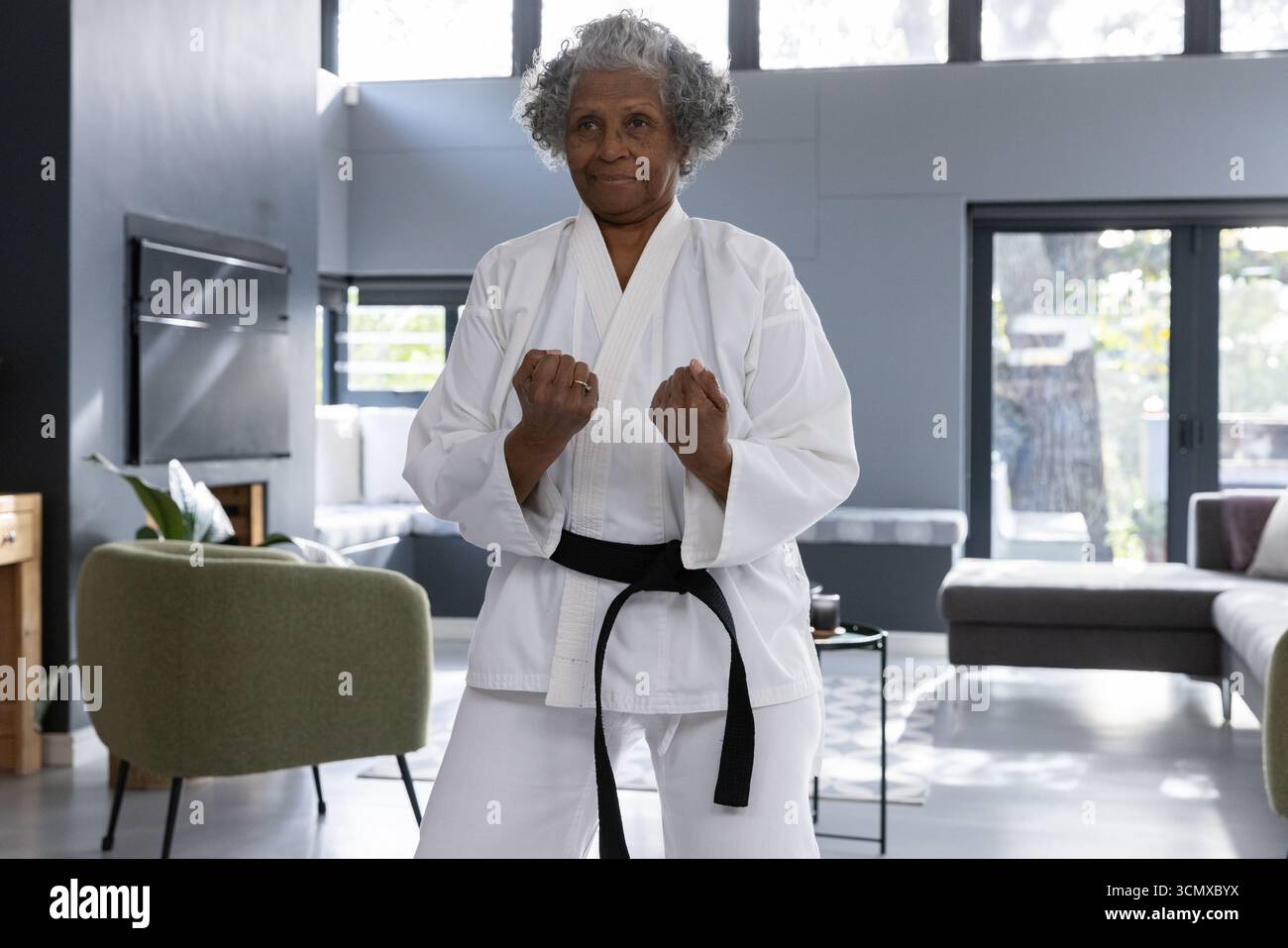 Femme afro-américaine senior tenant une position de combat dans le salon portant karaté GI et ceinture Banque D'Images