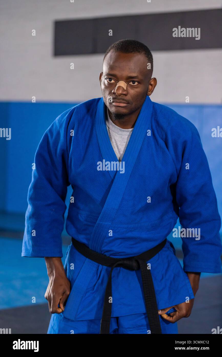 Homme afro-américain tenant la position de combat dans le dojo dans la ceinture noire GI bleue avec bandage nasal Banque D'Images Homme afro-américain tenant la position de combat dans le dojo dans la ceinture noire GI bleue avec bandage nasal Banque D'Images