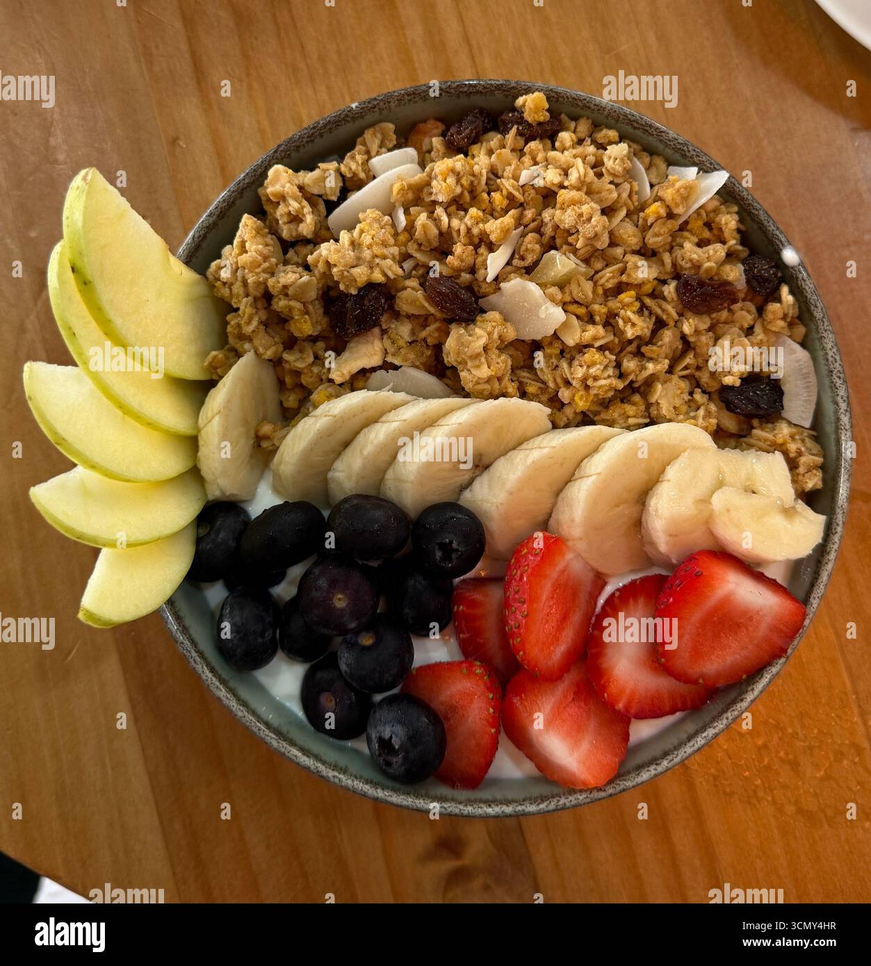 Bol de petit déjeuner sain avec granola, banane, fraises, bleuets et tranches de pommes sur yaourt, servi sur une table en bois. Banque D'Images