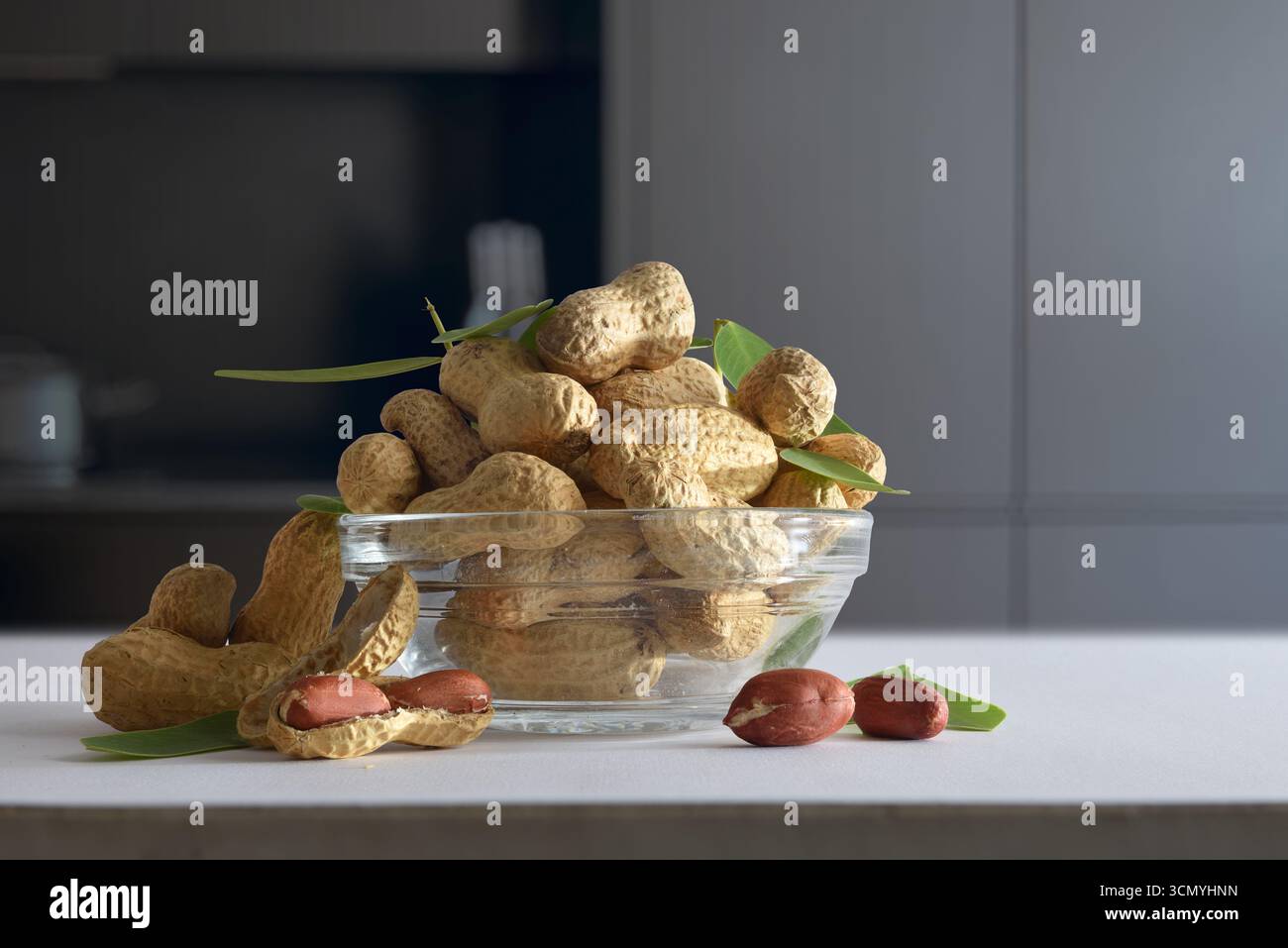 Détail d'un bol en verre avec un tas d'arachides décortiquées sur un banc de cuisine blanc avec des arachides ouvertes montrant les graines dans une cuisine sombre. Vue de face. Banque D'Images