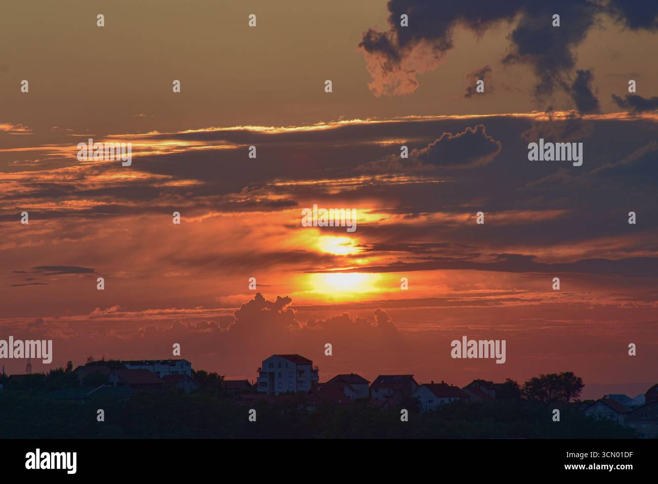 Ciel spectaculaire au coucher du soleil avec des tons gris et flamboyants qui brillent au-dessus des silhouettes de la ville lointaine reposant tranquillement sur une colline ombragée. Banque D'Images