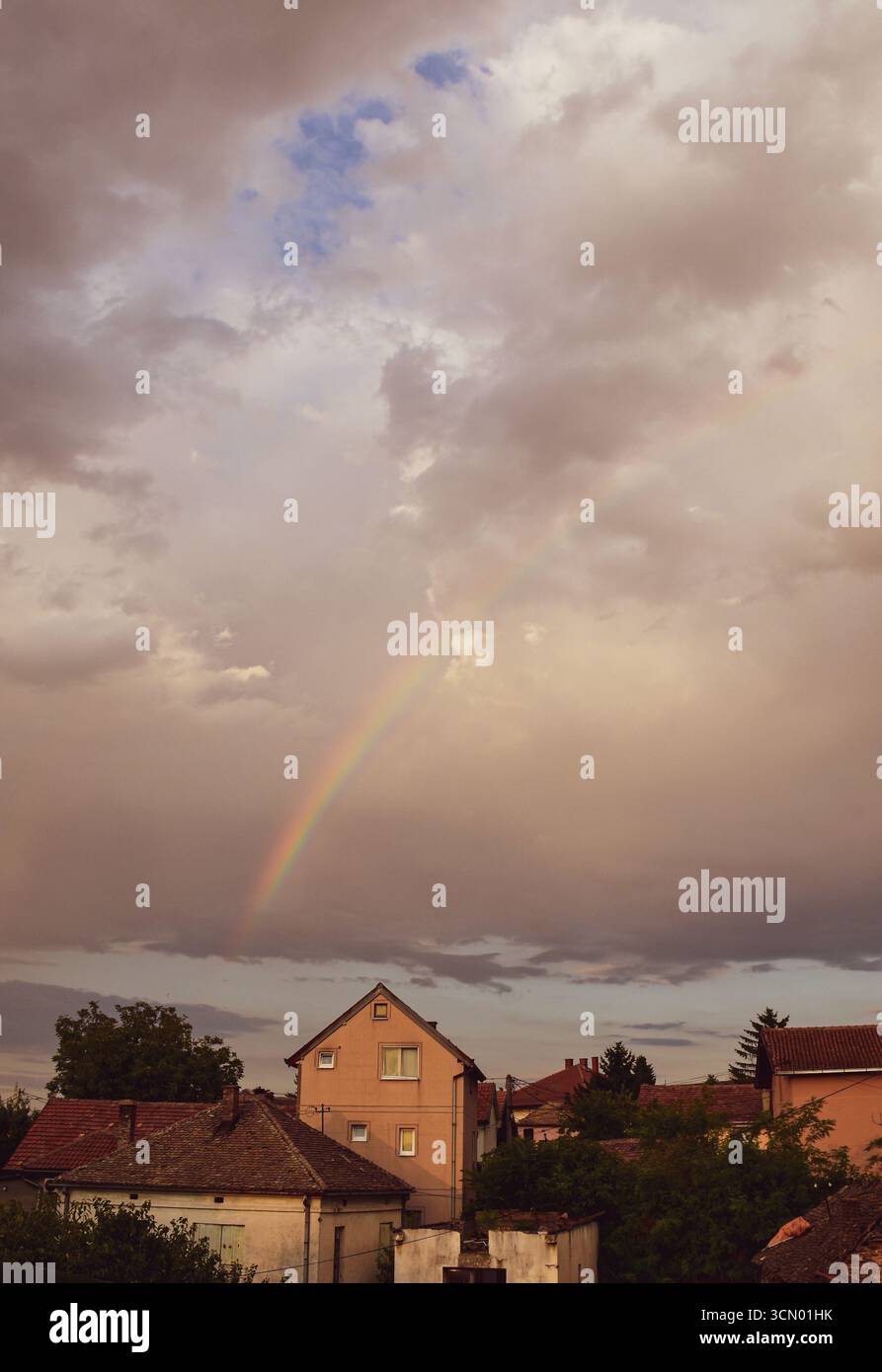 Un arc-en-ciel coloré au-dessus des nuages gris brille sur les maisons ensoleillées en contrebas, mêlant ciel orageux, lumière chaude et charme urbain. Banque D'Images