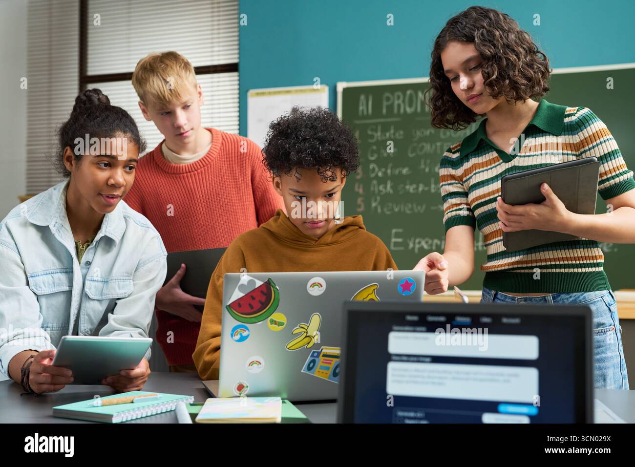 Groupe d'adolescents multiethniques collaborant sur un projet numérique en classe, en utilisant un ordinateur portable et des tablettes, debout et assis autour du bureau, axé sur la technologie et le travail d'équipe Banque D'Images