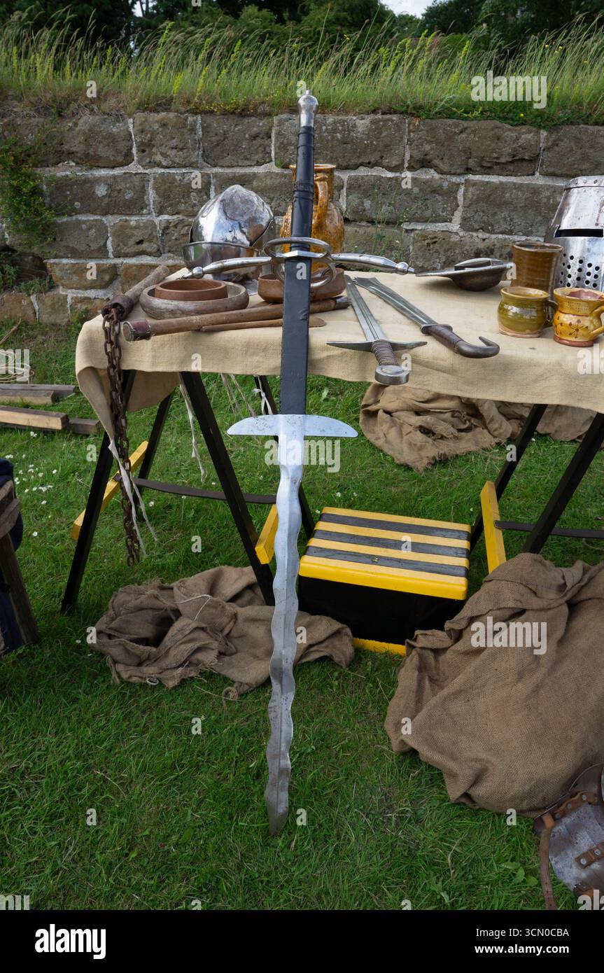 Une grande épée à deux mains avec une lame ondulée est appuyée contre une table d'objets médiévaux. Banque D'Images