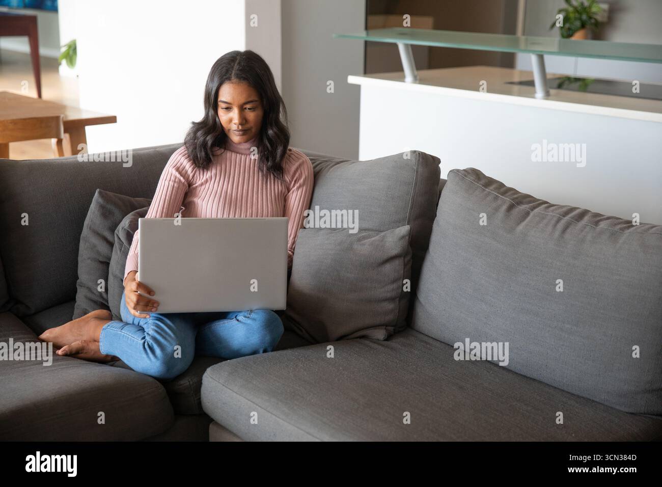 Femme assise à jambes croisées sur le canapé gris à l'aide d'un ordinateur portable argenté dans le salon, espace de copie Banque D'Images Femme assise à jambes croisées sur le canapé gris à l'aide d'un ordinateur portable argenté dans le salon, espace de copie Banque D'Images