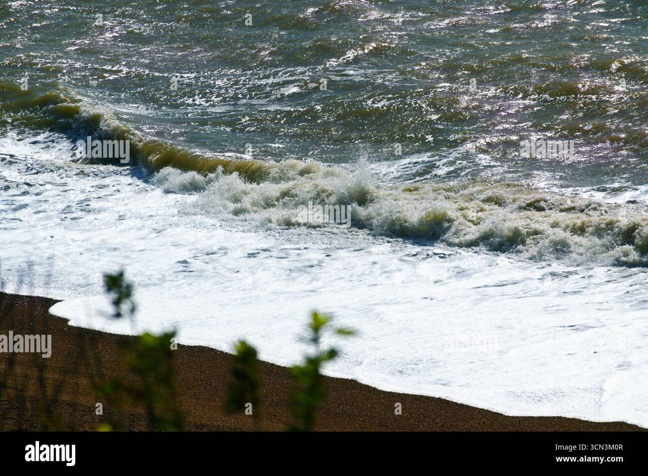 Une grosse vague s'écrase sur la plage de galets, créant une ligne de mousse blanche brillante contre l'eau sombre et bouillonnante de l'océan par une journée ensoleillée et venteuse Banque D'Images