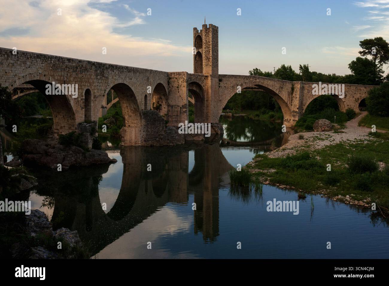 Par une chaude soirée de juin, la paisible rivière Fluvià reflète les arches inégales et la tour de défense centrale du Viejo médiéval reconstruit, Pont Vell ou Vieux Pont, construit sur les rochers du lit de la rivière à Besalú, Gérone, Catalogne, Espagne. La structure fortifiée inclinée du 11ème siècle ce, d'environ 105m ou 344 pieds de long, est un monument emblématique catalan et un emblème populaire de la ville antique. Banque D'Images