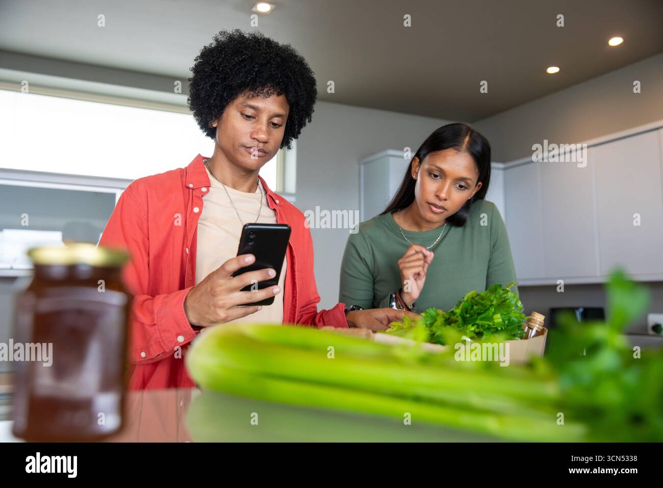 Couple diversifié inspectant les herbes fraîches dans une caisse en bois, défilant smartphone à l'îlot de cuisine Banque D'Images Couple diversifié inspectant les herbes fraîches dans une caisse en bois, défilant smartphone à l'îlot de cuisine Banque D'Images